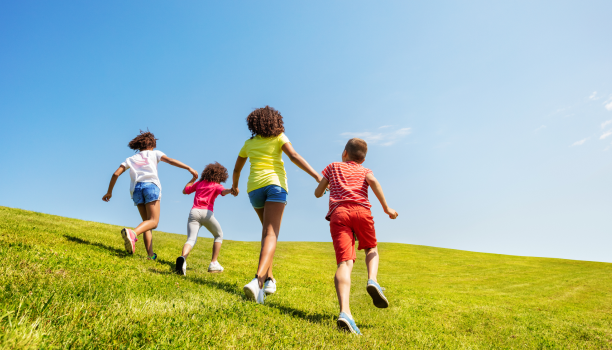 A group of children are shown from behind holding hands and skipping in on a green hill. 